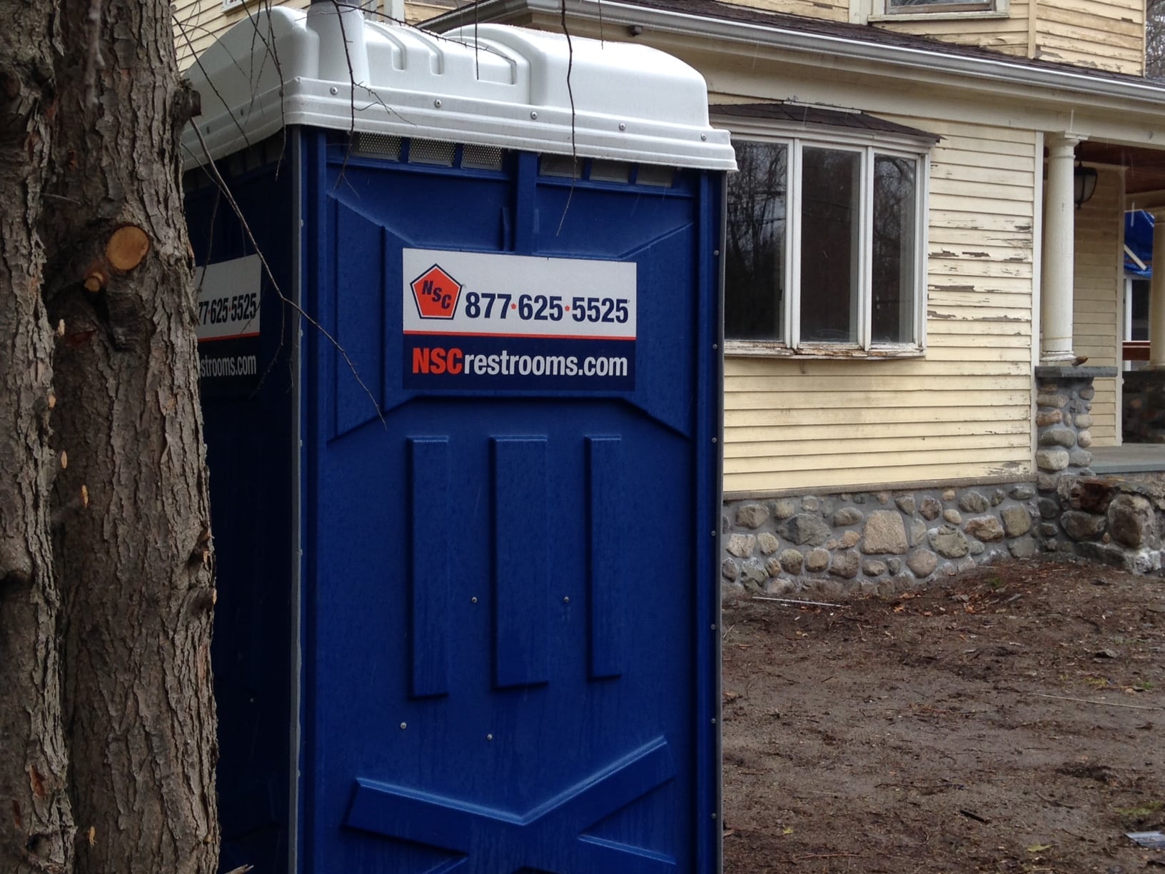 The deep blue of this port-a-potty brings to mind the clear sky of a beautiful spring day, while the weathered yellow clapboards of the house evoke the weak sunshine common early in the season.