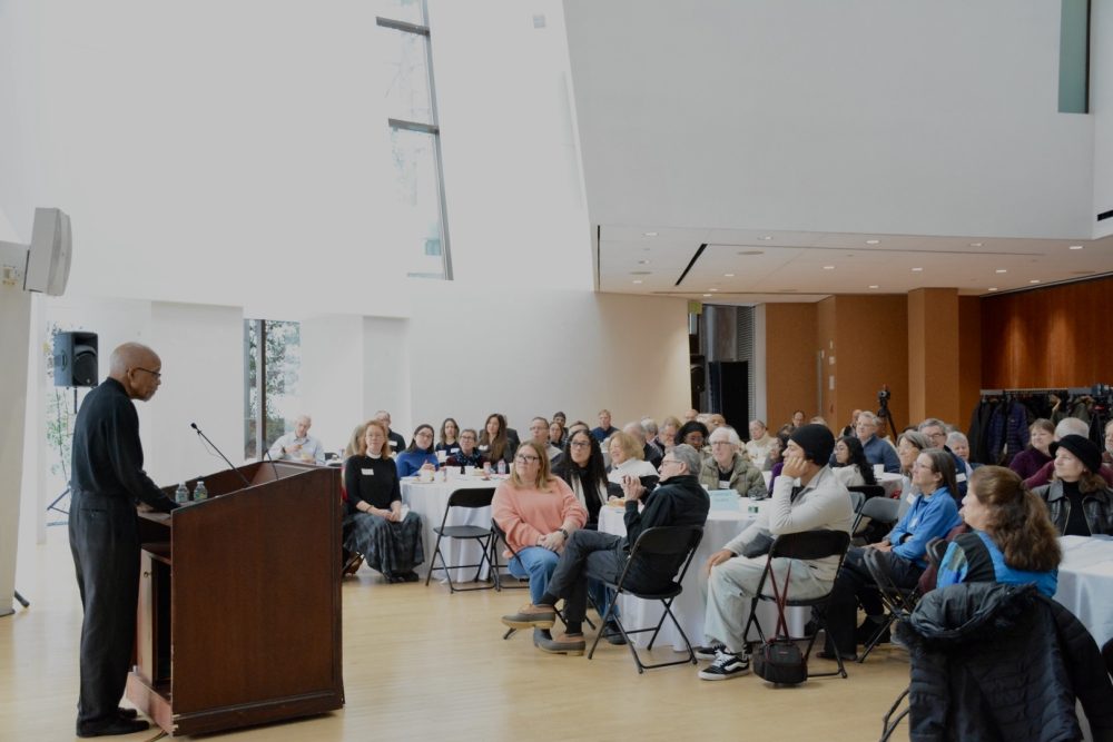 A group of people gathered in Tishman Commons listen to a man speaking at the podium.